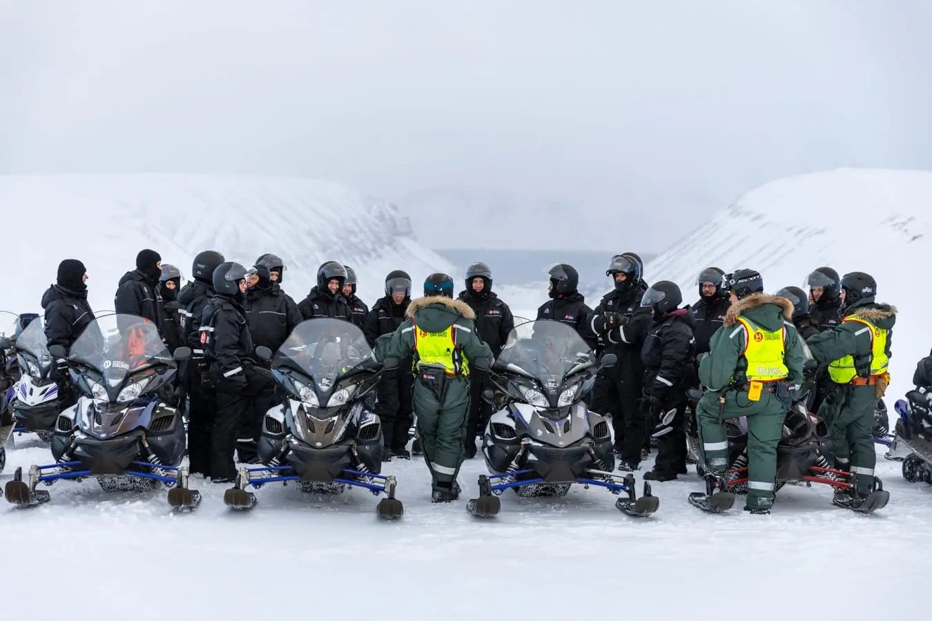 En stor gruppe i svarte dresser og hjelmer samlet ved snøscootere med guider i grønt, foran snødekte fjell