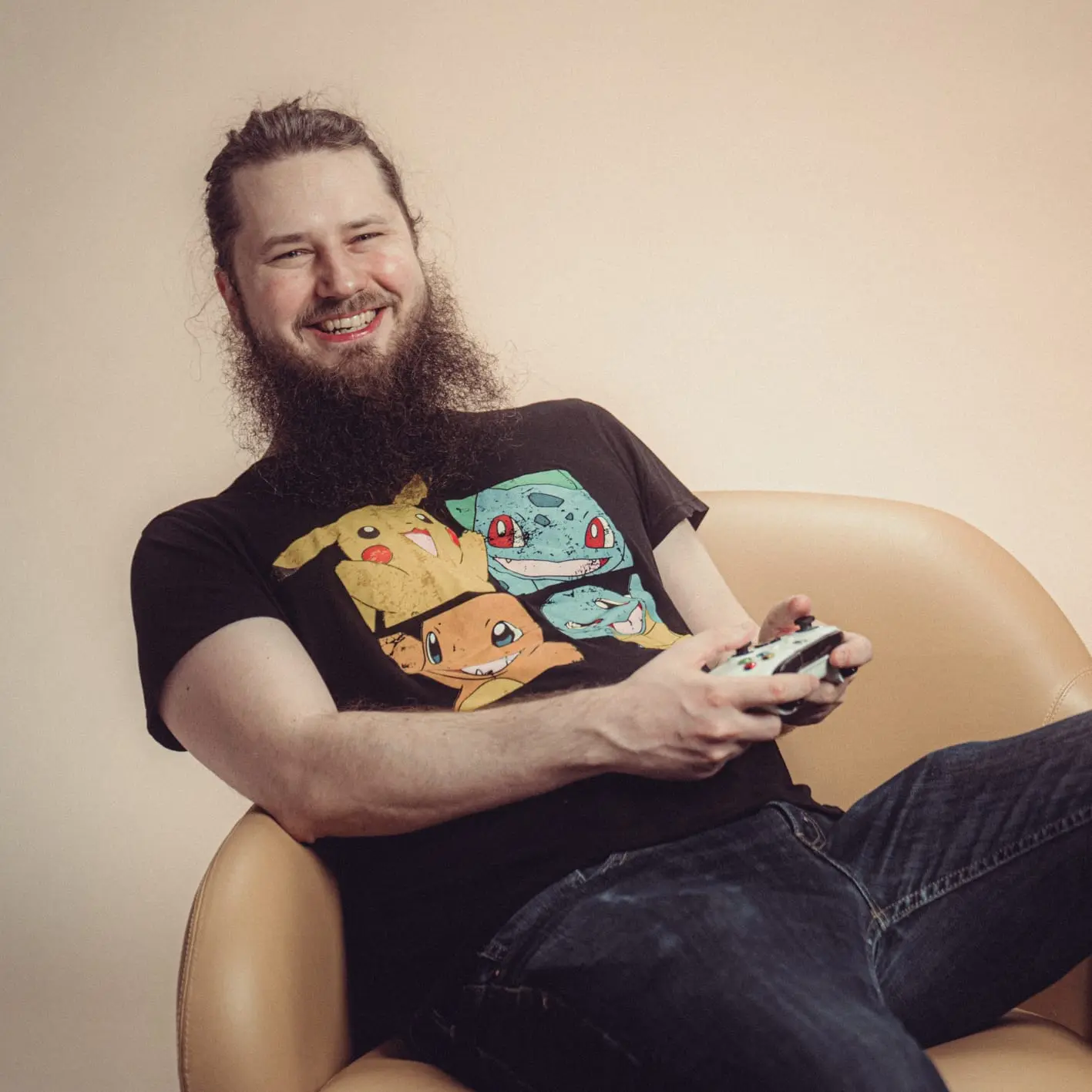 En smilende mann med langt skjegg og Pokémon-t-skjorte sitter i en lenestol med en spillkontroller