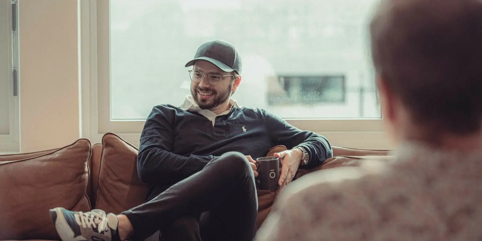 En smilende mann i caps sitter i en skinnsofa med en kaffekopp og prater med en kollega
