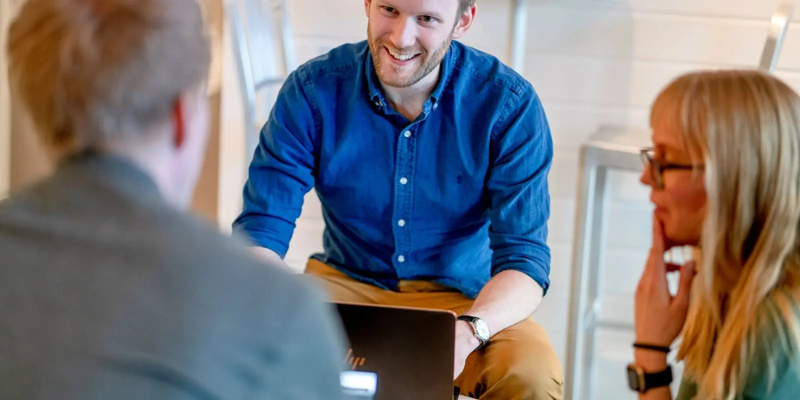 En smilende mann i blå skjorte diskuterer med to kolleger rundt en bærbar PC