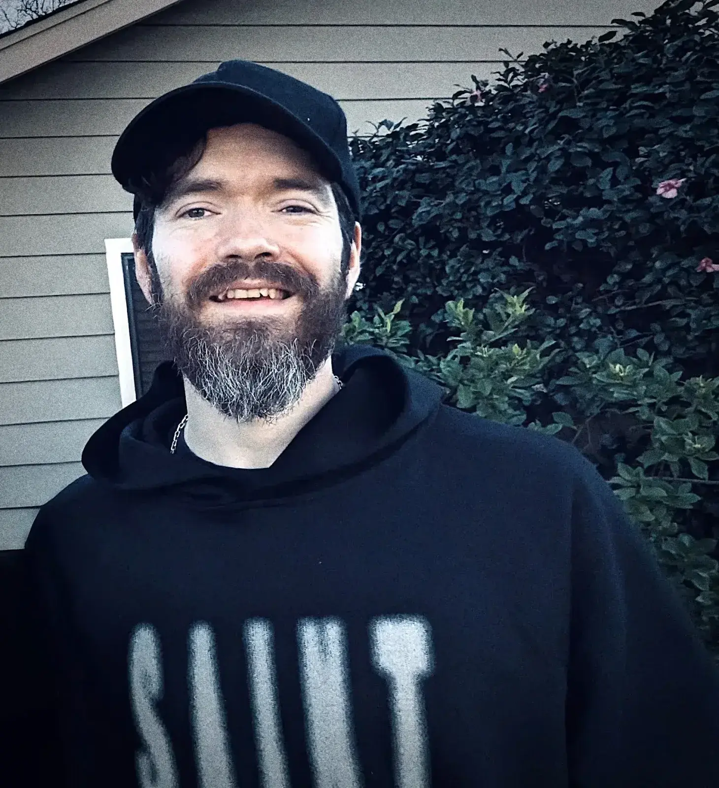 En smilende mann med skjegg, svart caps og hettegenser foran et hus