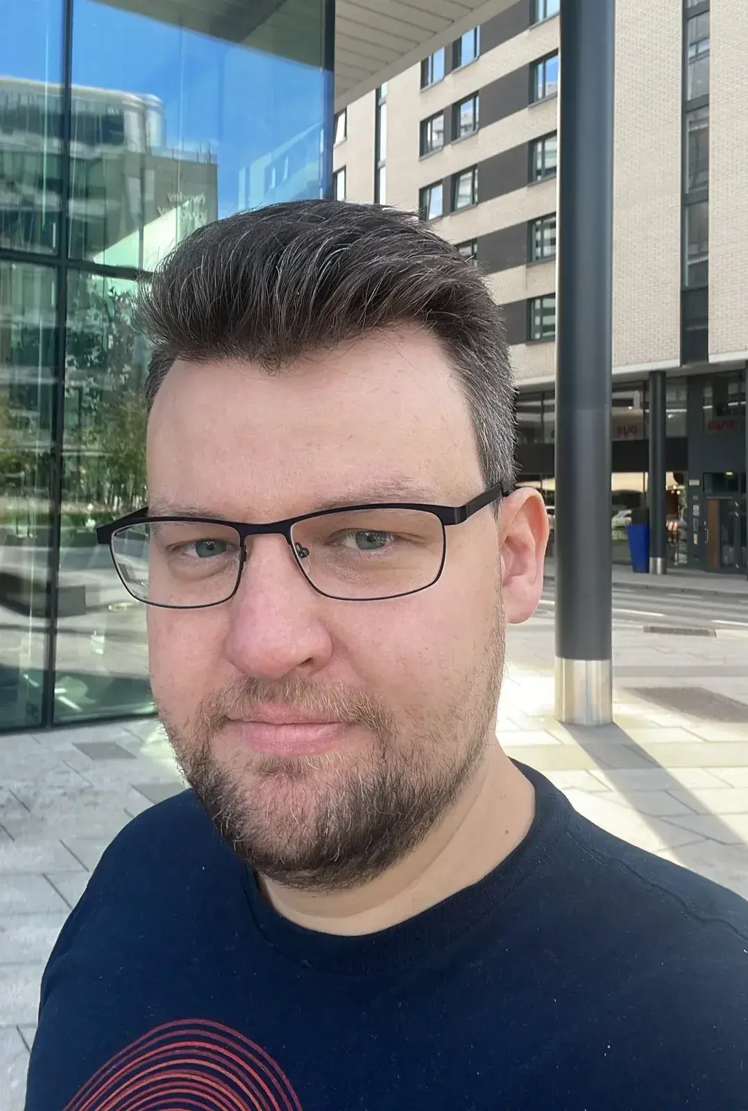 Selfie av en mann med briller og skjegg foran et bygg i byen