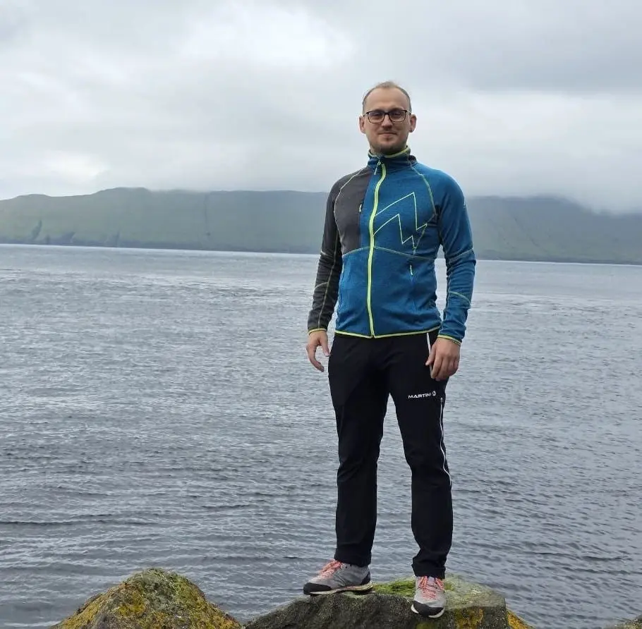 En mann med briller står på en stein ved sjøen med grønne fjell i bakgrunnen