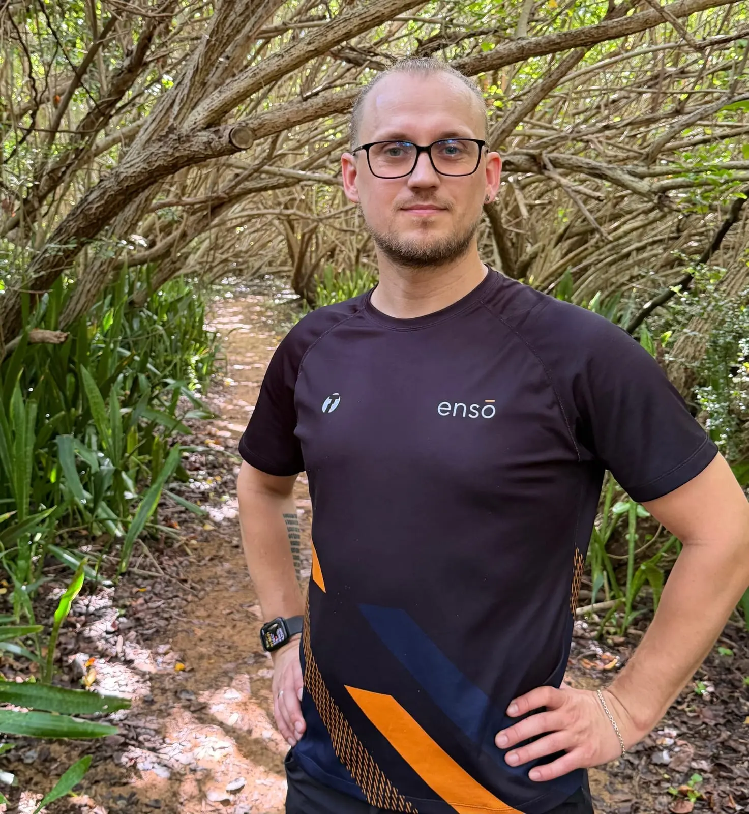 En mann med briller i Ensō-treningstrøye står på en sti i en skog