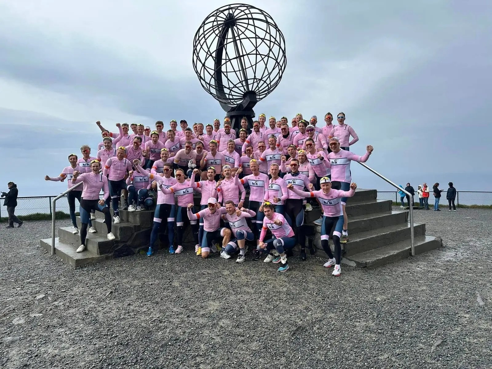 Syklister i rosa klubbdrakter jubler foran globusmonumentet på Nordkapp