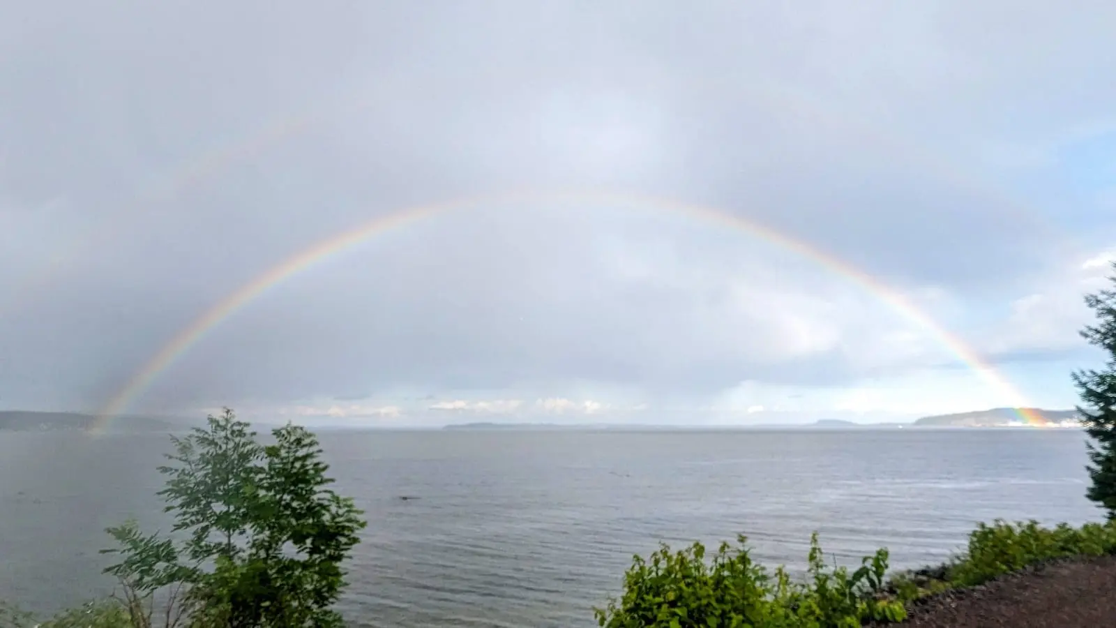 Dobbel regnbue over en bred fjord en grå sommerdag, med busker i forgrunnen
