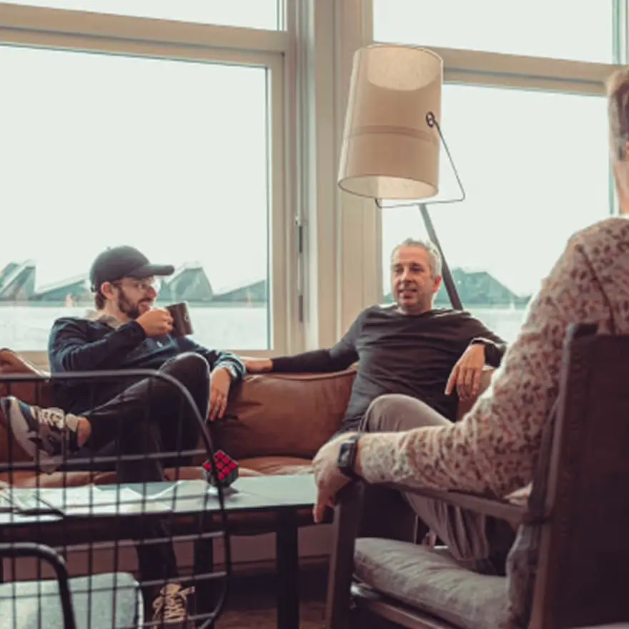 Kolleger med kaffe i sofakroken ved de store vinduene på kontoret.